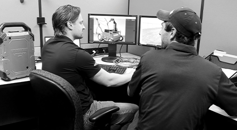 Two engineers in a Vanguard product laboratory talk to each other while sitting in front of two computer monitors. A Vanguard battery pack is in the background.