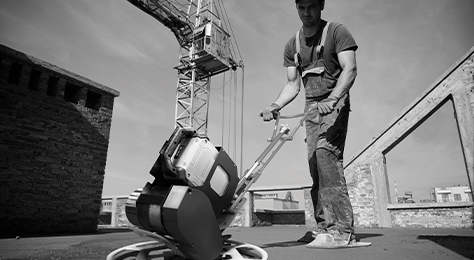 A construction worker uses a battery-powered walk-behind concrete trowel at an outdoor jobsite. A crane is in the background.